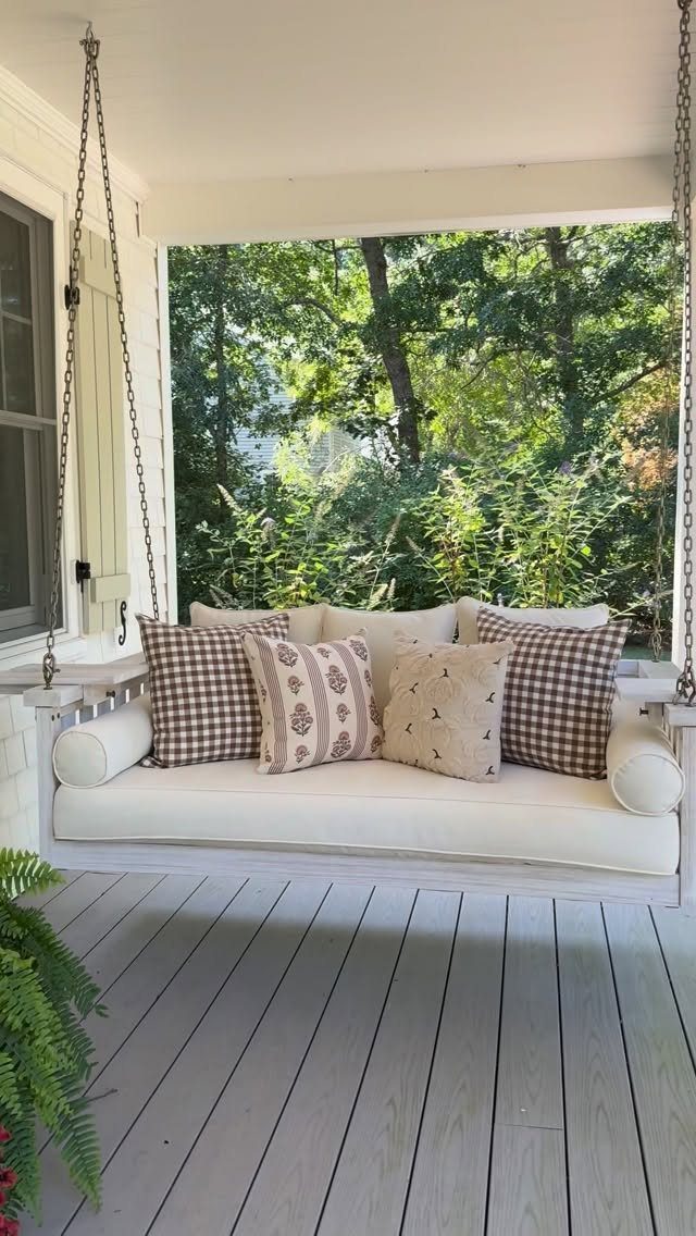 A white hanging porch swing with decorative pillows sits on a wooden porch overlooking green trees.