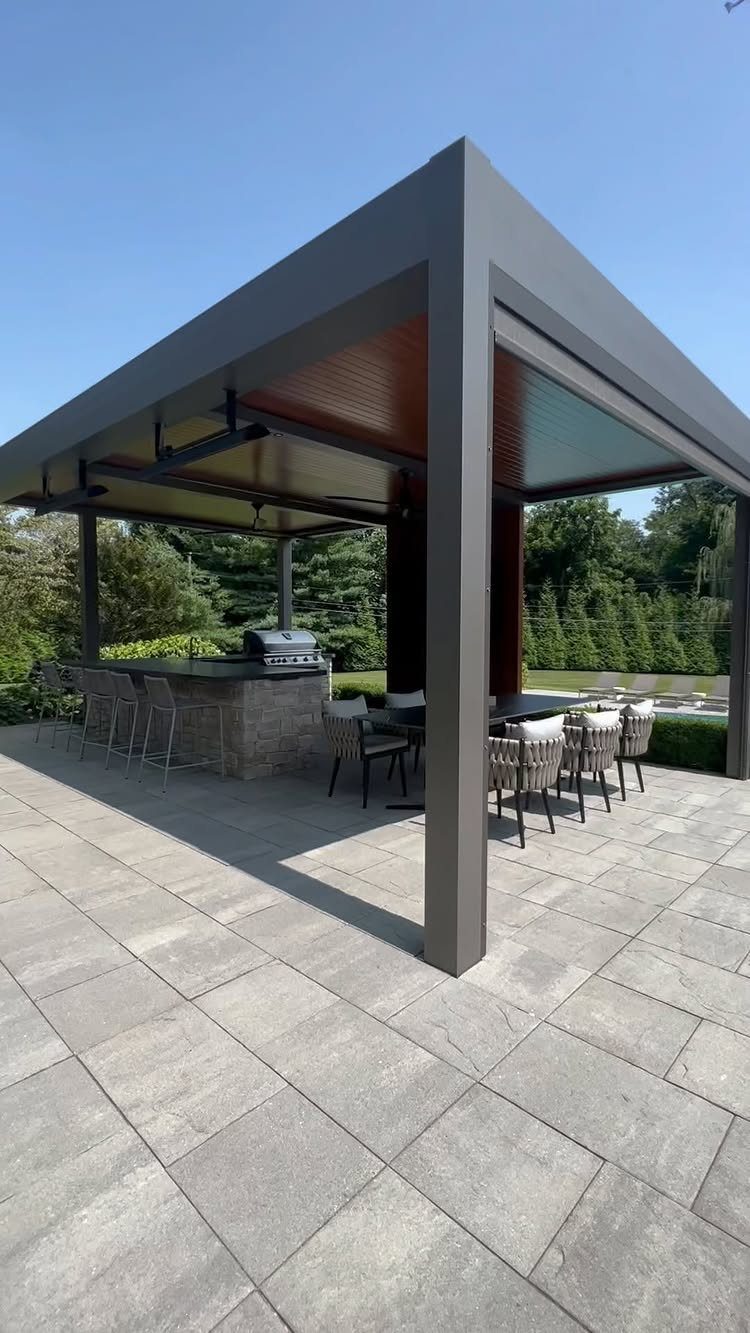 Modern outdoor patio with a dark pergola, stone kitchen bar, and dining set on grey tiles.