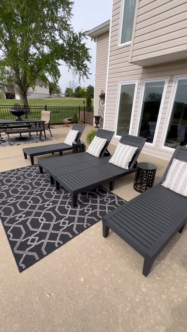 Four dark grey lounge chairs with patterned pillows sit on a patterned rug on a concrete patio.
