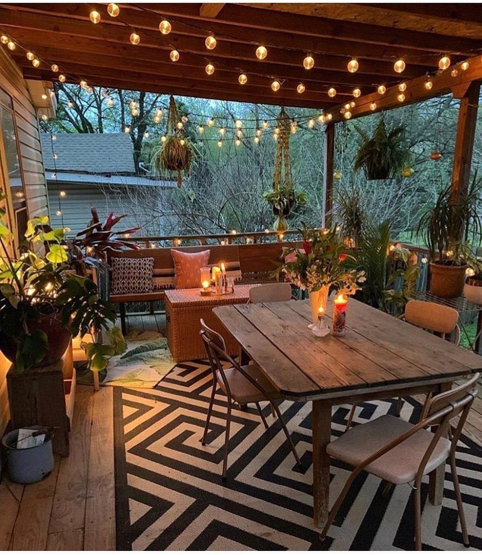 A decorated wooden back porch with string lights, dining furniture, and lush green plants.