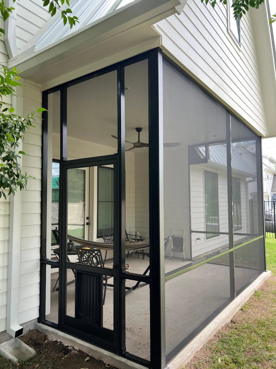 A black-framed screened-in porch attached to a white siding house with patio furniture inside.