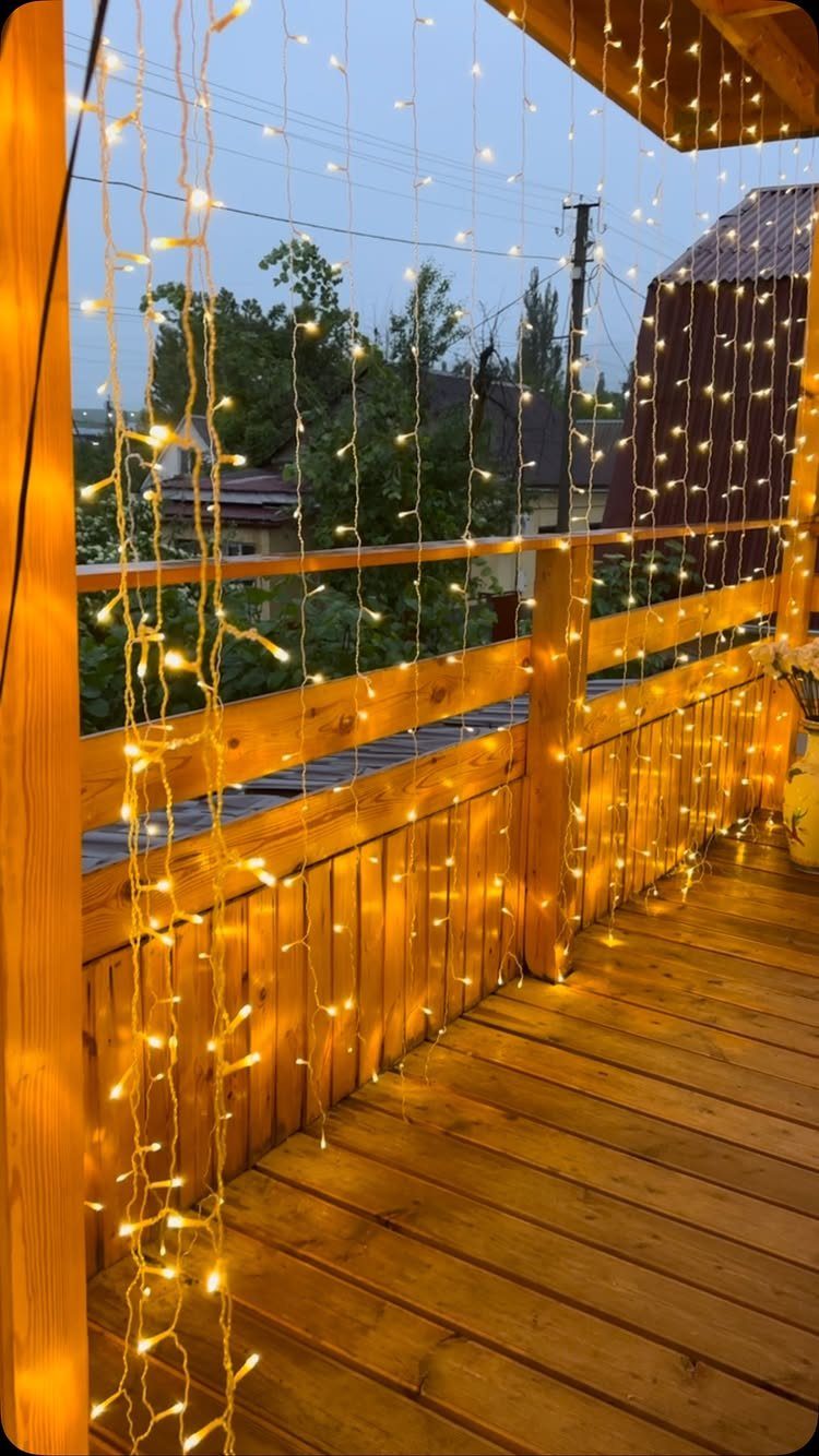 Warm string lights hanging vertically on a wooden back porch balcony overlooking green trees at dusk.