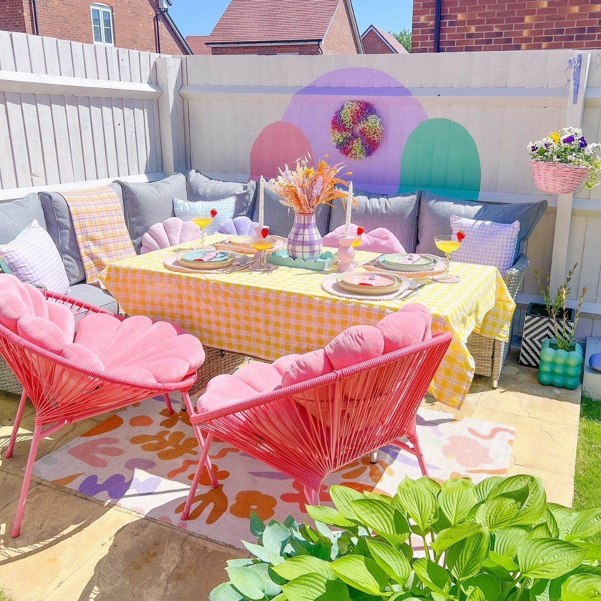Pink petal chairs and colorful cushions surround a yellow checkered dining table in a sunny backyard.