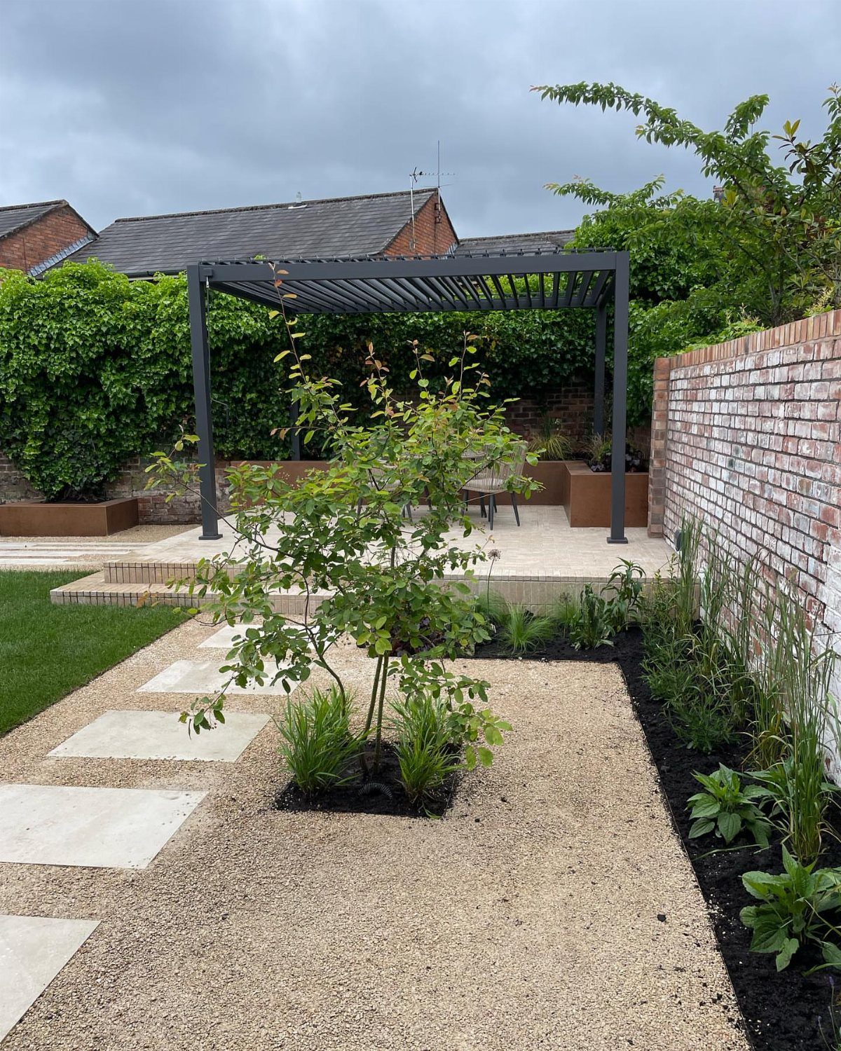 Light gravel pathway with large stone pavers leading toward a modern dark pergola in a garden.