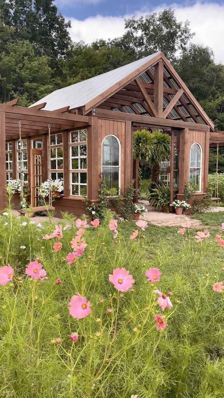 Wooden greenhouse structure with large glass windows surrounded by pink cosmos flowers and green trees.
