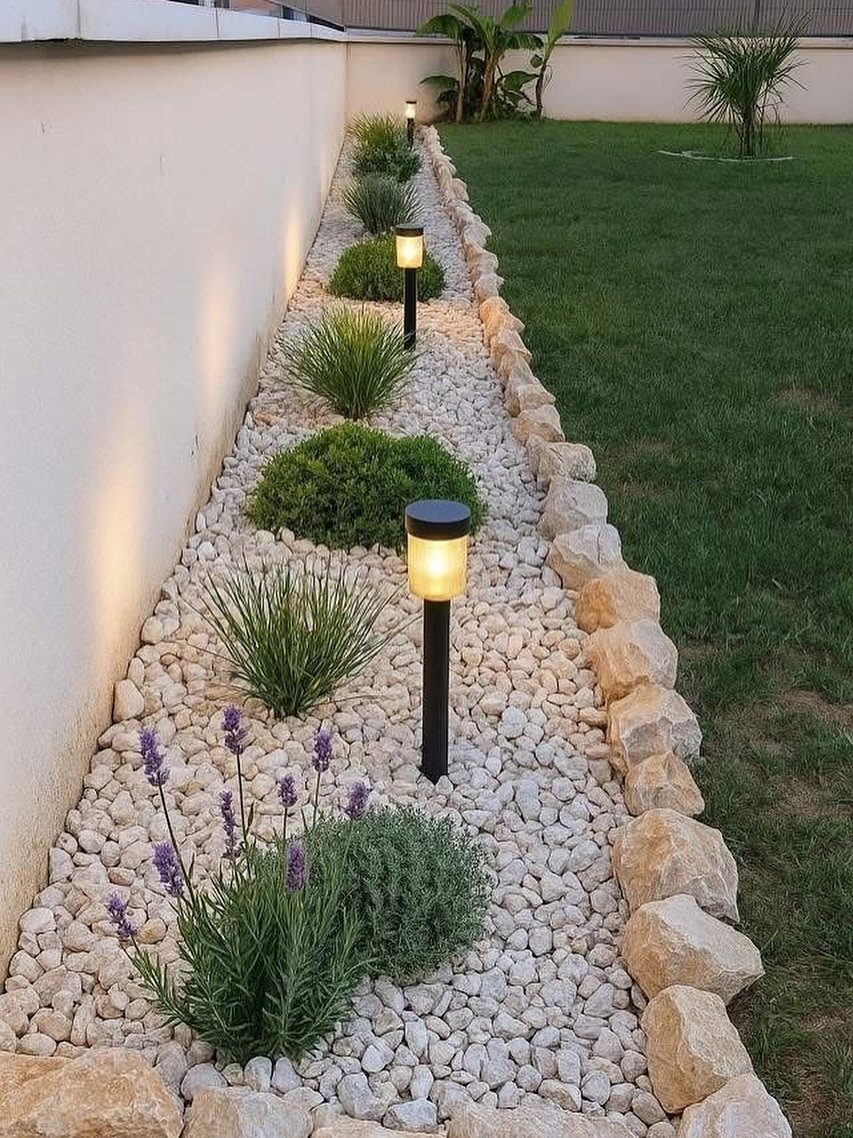Small green shrubs and solar lights planted in a white pebble garden bed lined with stones.