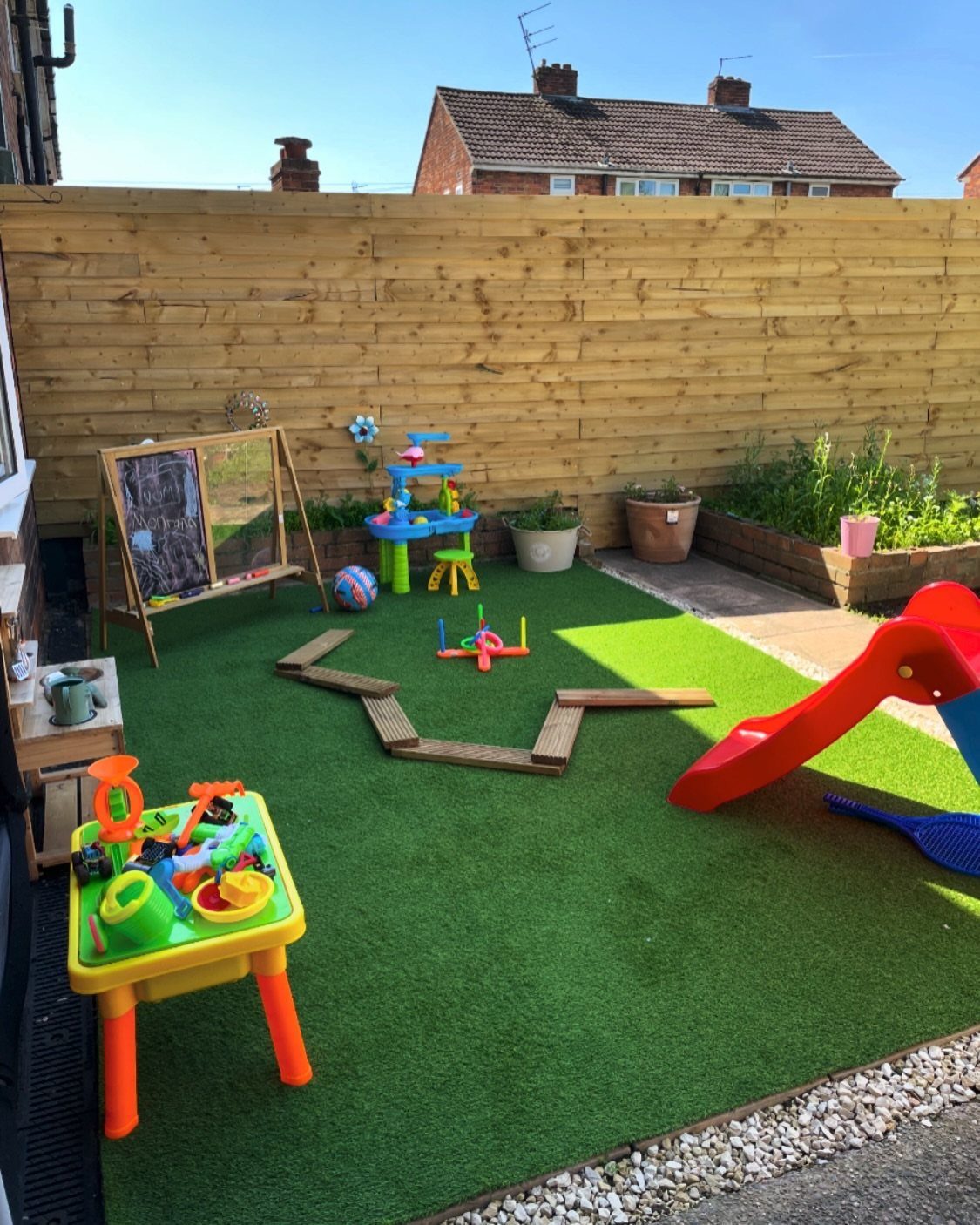 Colorful children's toys and a red slide on green artificial grass in a backyard.