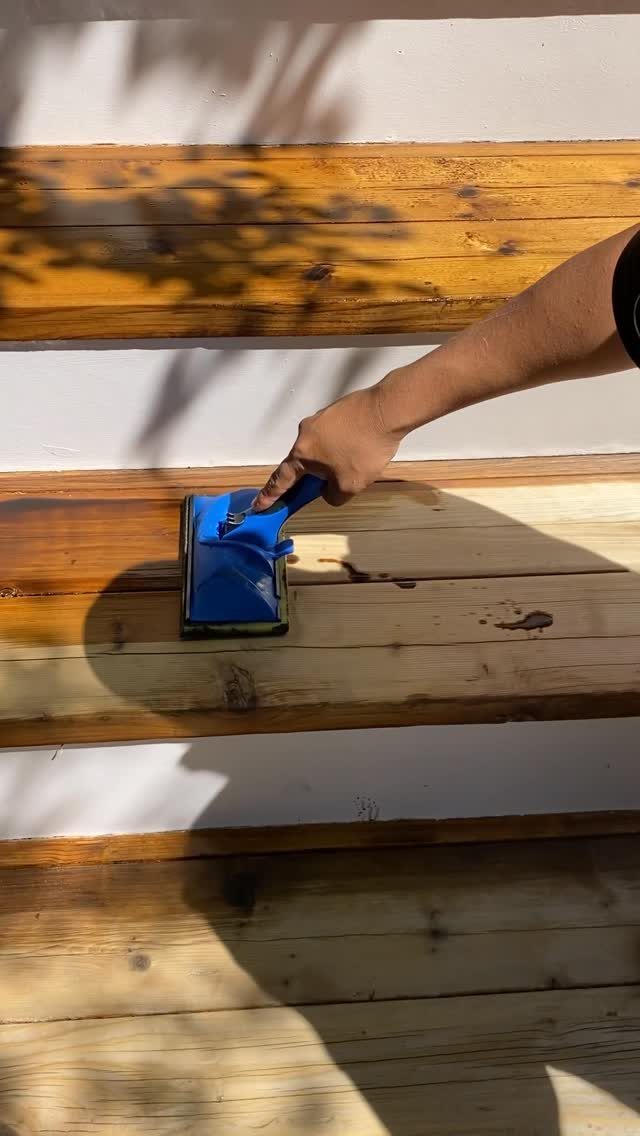A hand applying wood stain to cedar planks using a blue applicator tool.