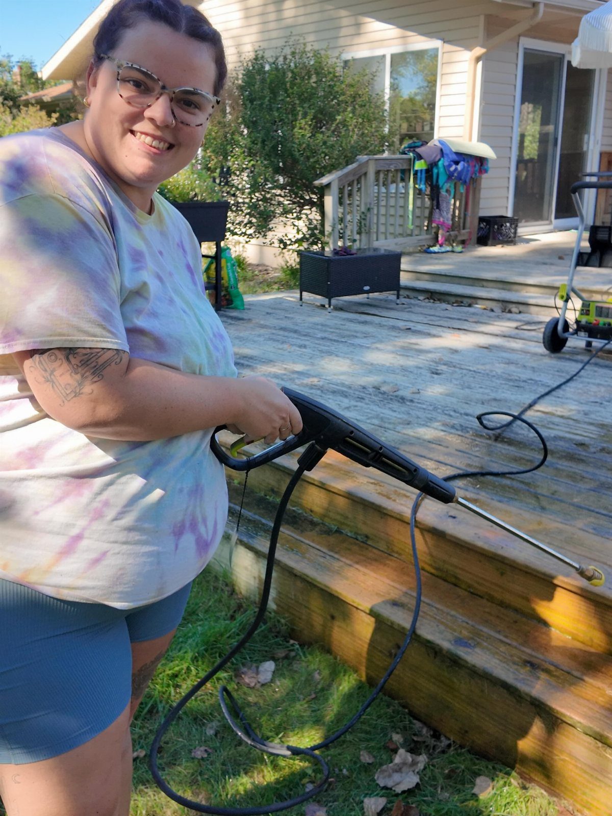 A smiling woman uses a pressure washer to clean a wooden outdoor deck.