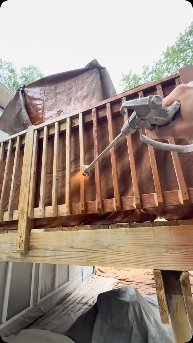 A hand holds a spray gun applying liquid to a wooden deck railing outdoors.