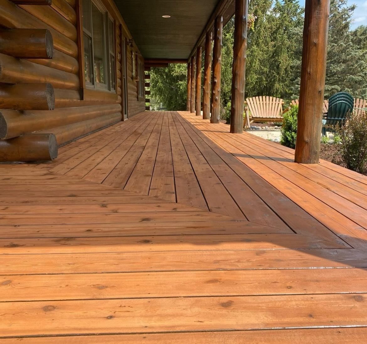 A wide wooden deck with a reddish-brown transparent stain and chevron pattern attached to a log cabin.
