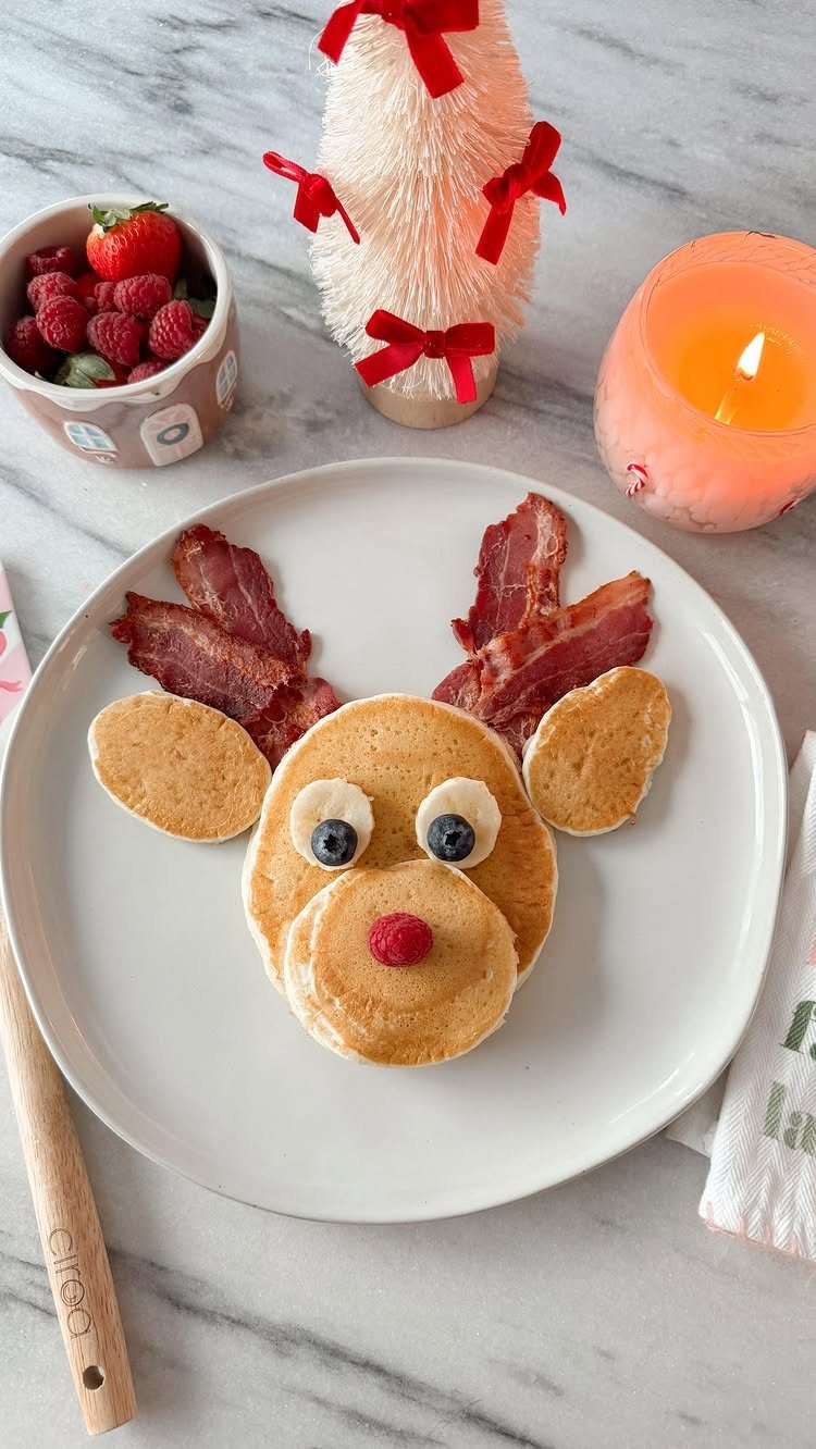 Reindeer-shaped pancakes with bacon antlers and fruit features on a white plate.