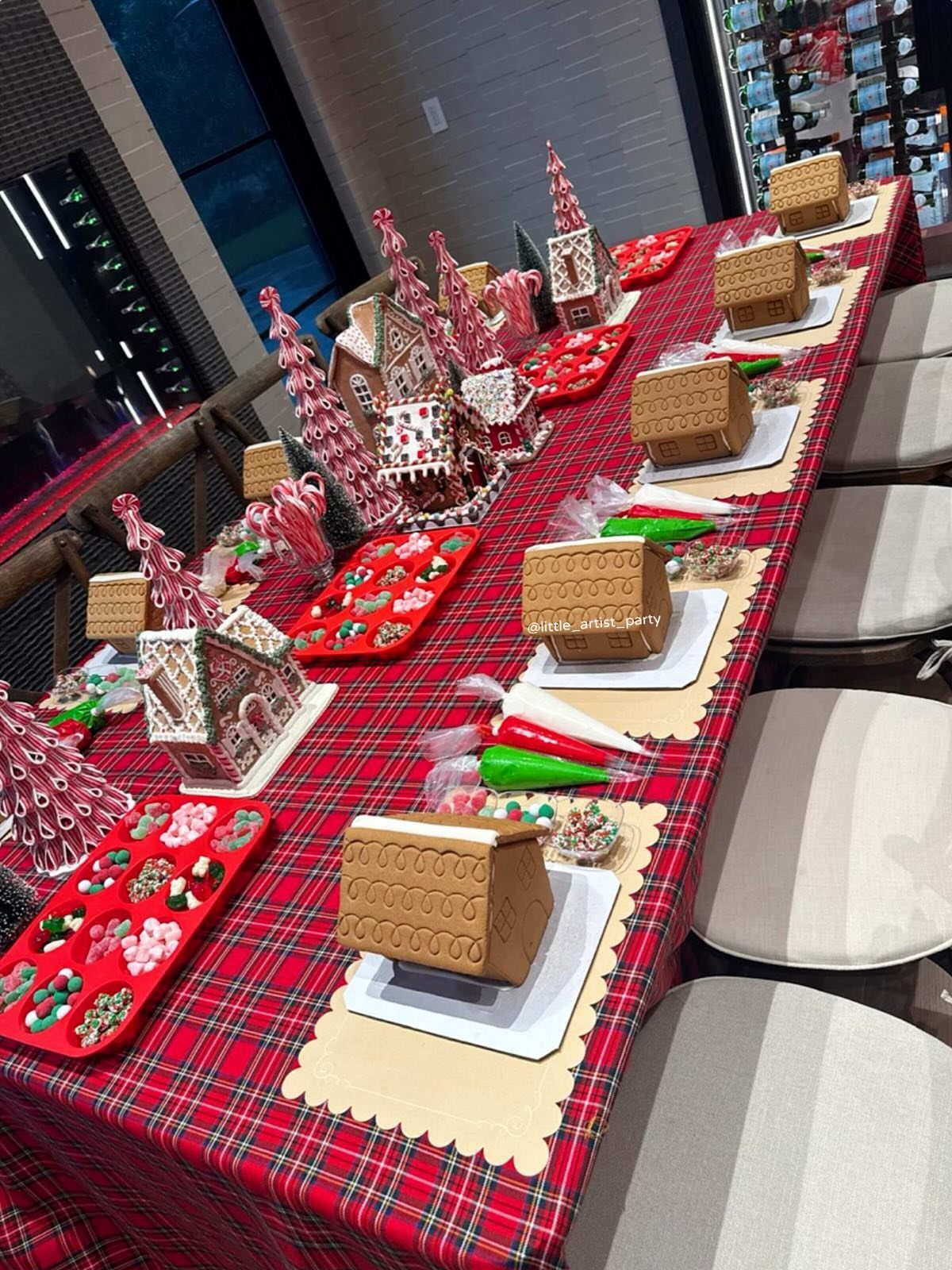 Gingerbread houses and decorating supplies arranged on a long red plaid tablecloth.