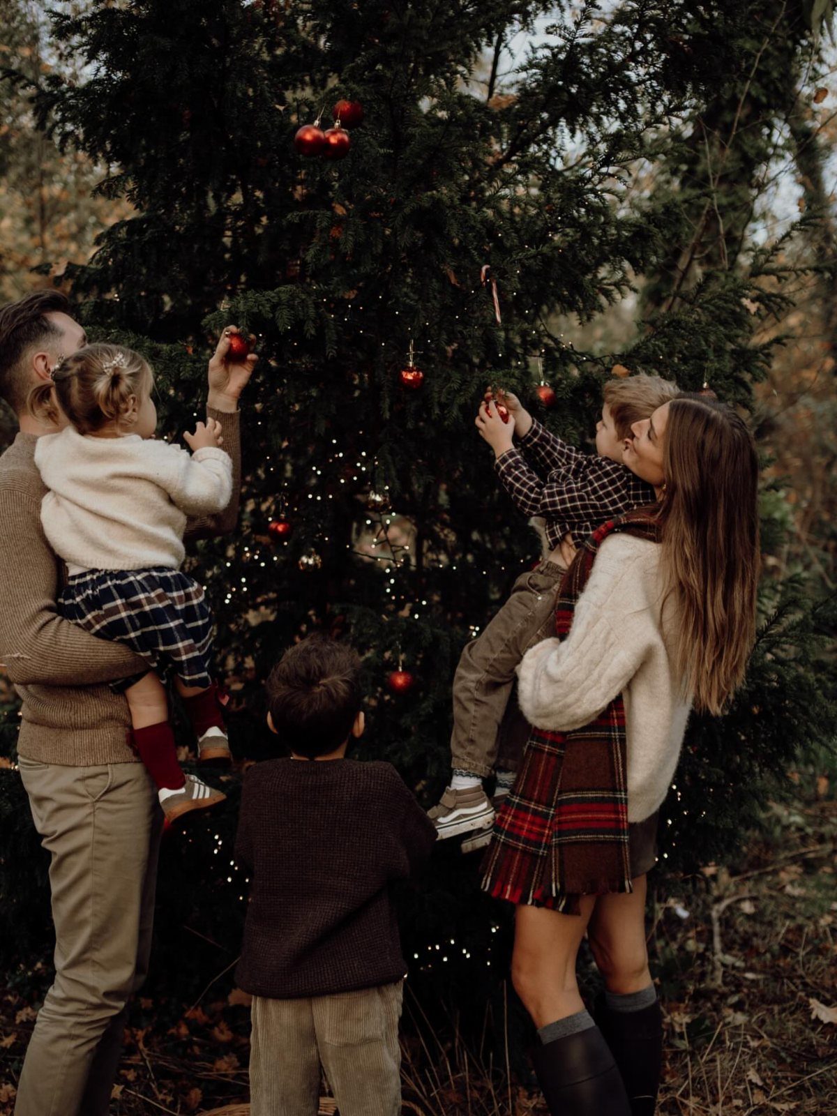 A family decorating a large outdoor Christmas tree with red ornaments in a forest.