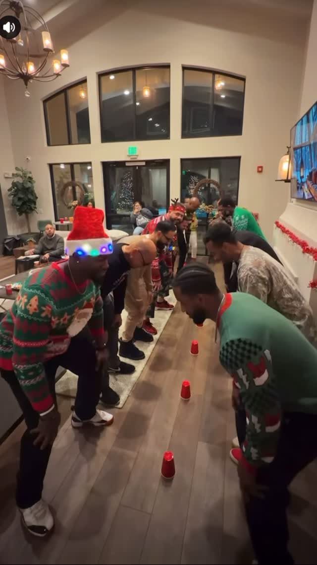 Men in Christmas sweaters playing a cup game on a wooden floor indoors.