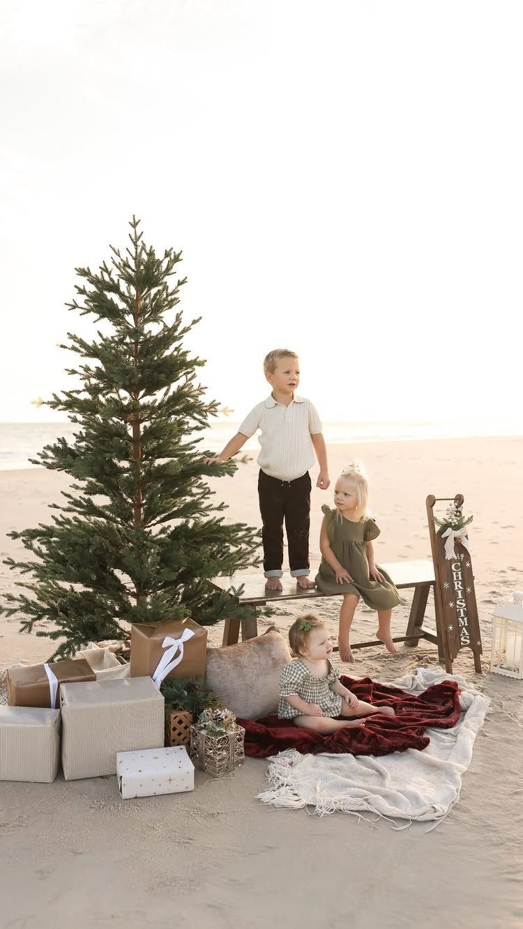 Three children posing on a beach with a small Christmas tree and gifts.