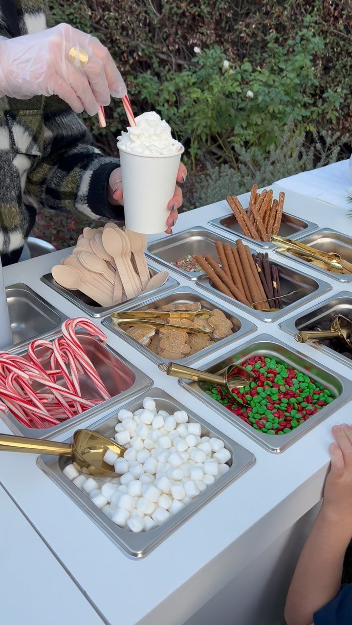 A person holding a whipped cream topped drink near trays of festive hot chocolate toppings.