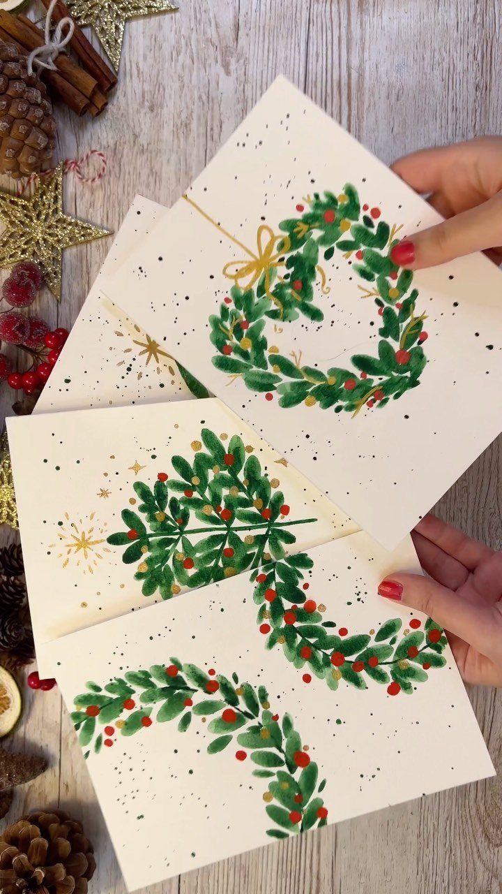 Hands holding handmade Christmas cards decorated with green watercolor wreaths and red berries on wood.