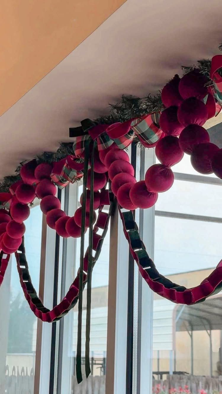 Red pom-pom garland with plaid ribbon hanging along a white ceiling near a window.