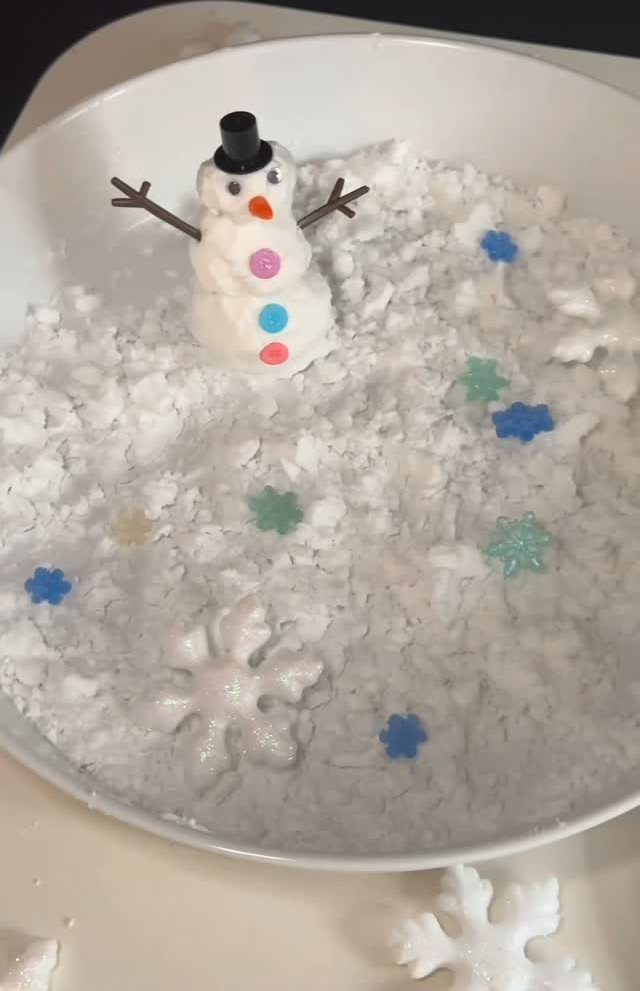 A small snowman figurine sitting in a white bowl of fluffy fake snow and snowflakes.