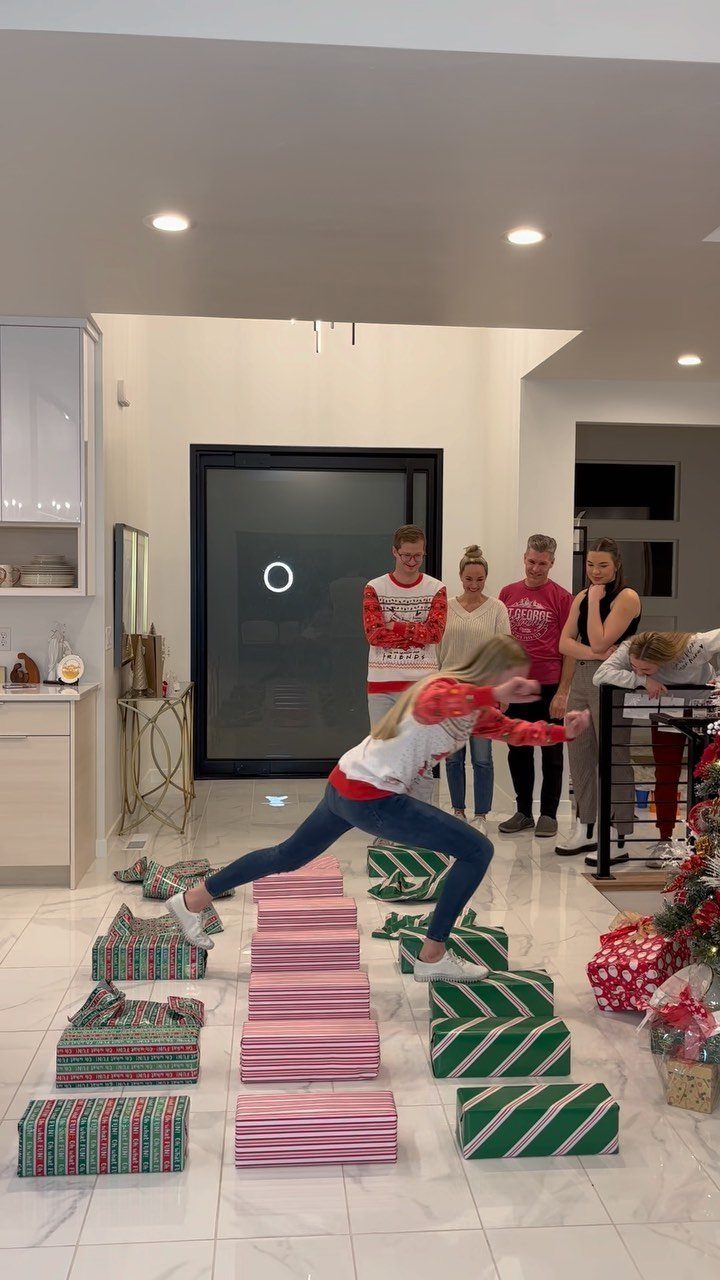 A young girl jumps over a row of pink Christmas presents in a brightly lit living room.