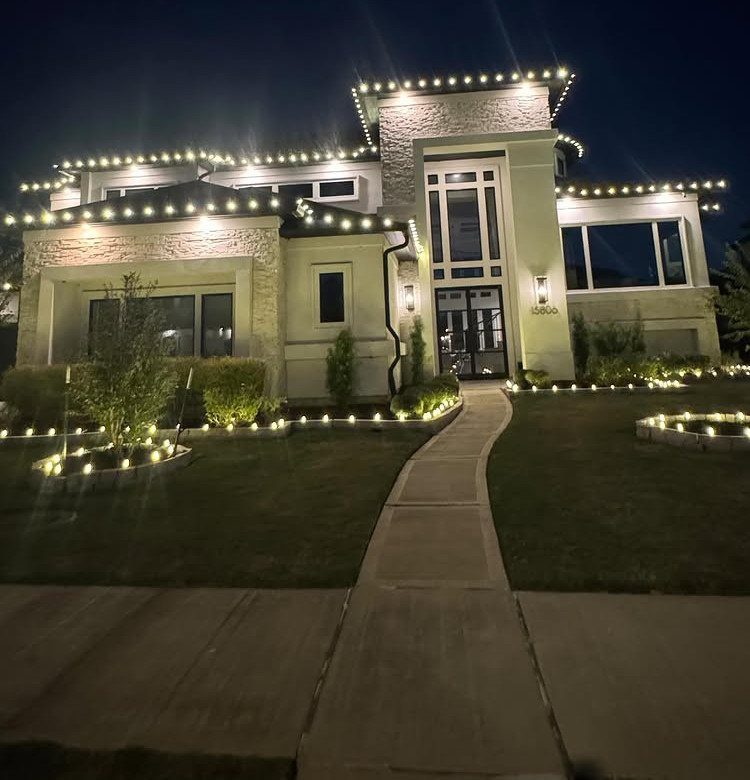 A large modern house decorated with warm white Christmas lights along the roof and garden edges.