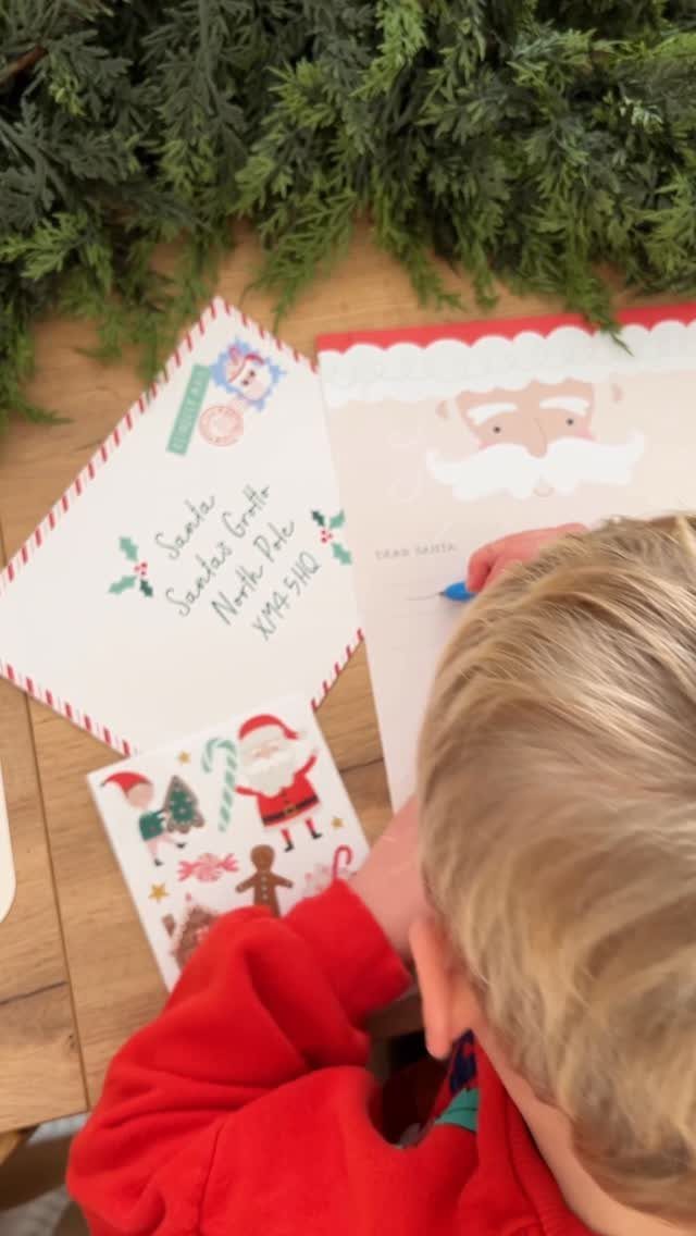 A child in a red sweater writing on a large Santa Claus themed paper on a wooden table.