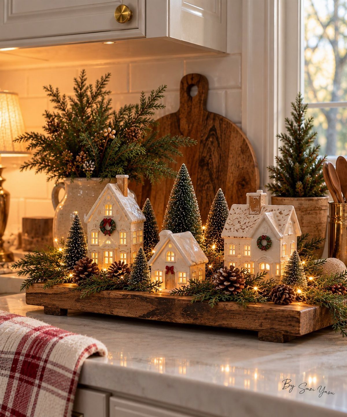 Ceramic miniature houses and evergreen trees arranged on a wooden tray with pinecones and greenery.