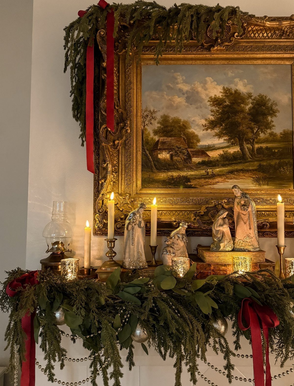 Antique figurines and lit candles arranged on a mantel with green garlands and red ribbons.