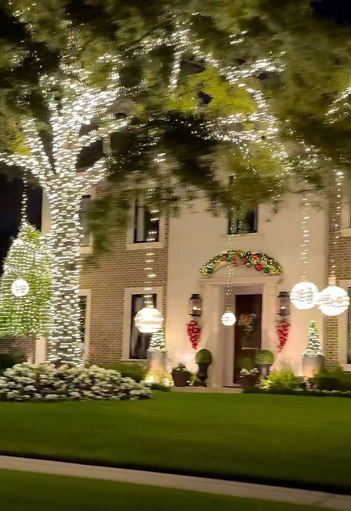 A large tree wrapped in white lights stands before a house decorated for Christmas.