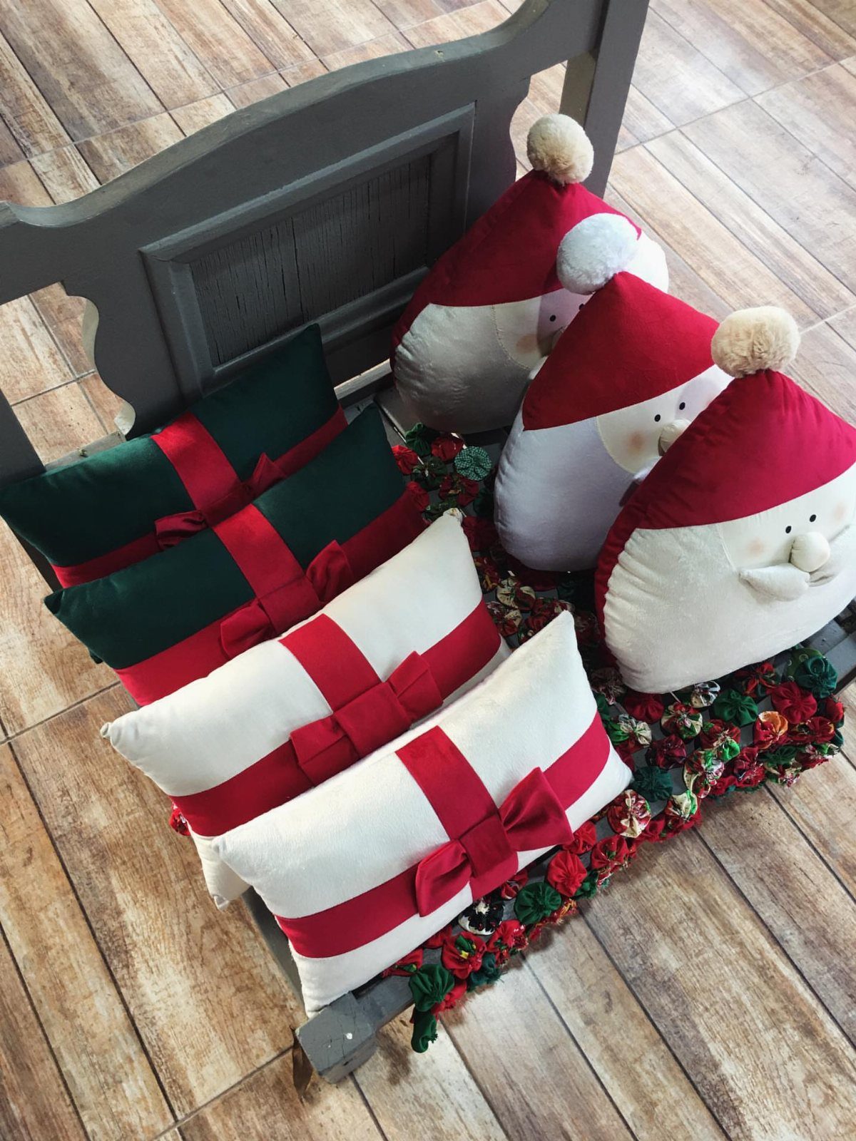 Decorative gift-shaped and Santa Claus pillows resting on a wooden bench with floral garland.