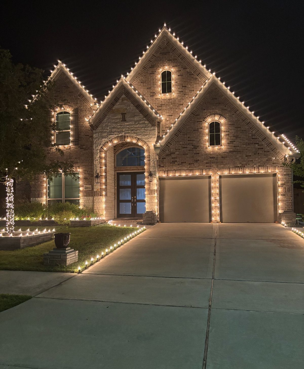 A brick house at night decorated with warm white Christmas lights along the roof and driveway.