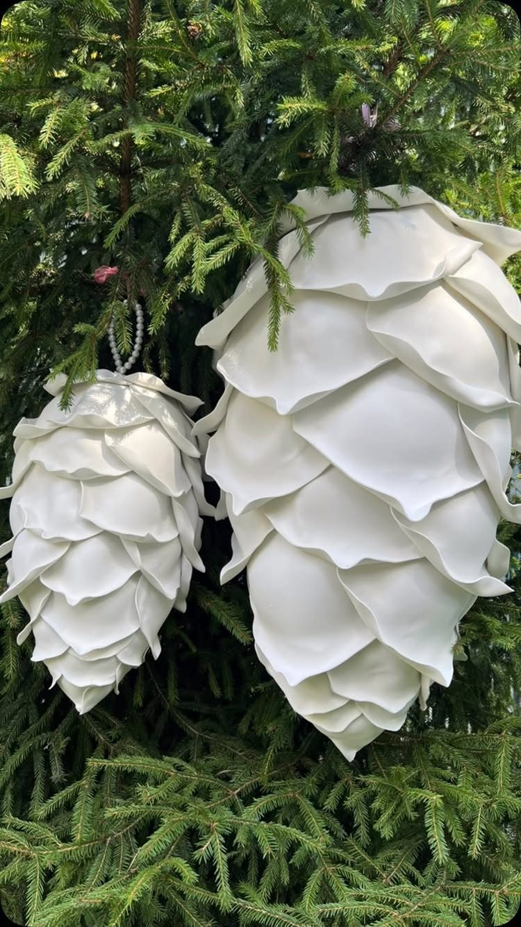 Two large white sculpted pinecone ornaments hanging from the branches of a green evergreen tree.
