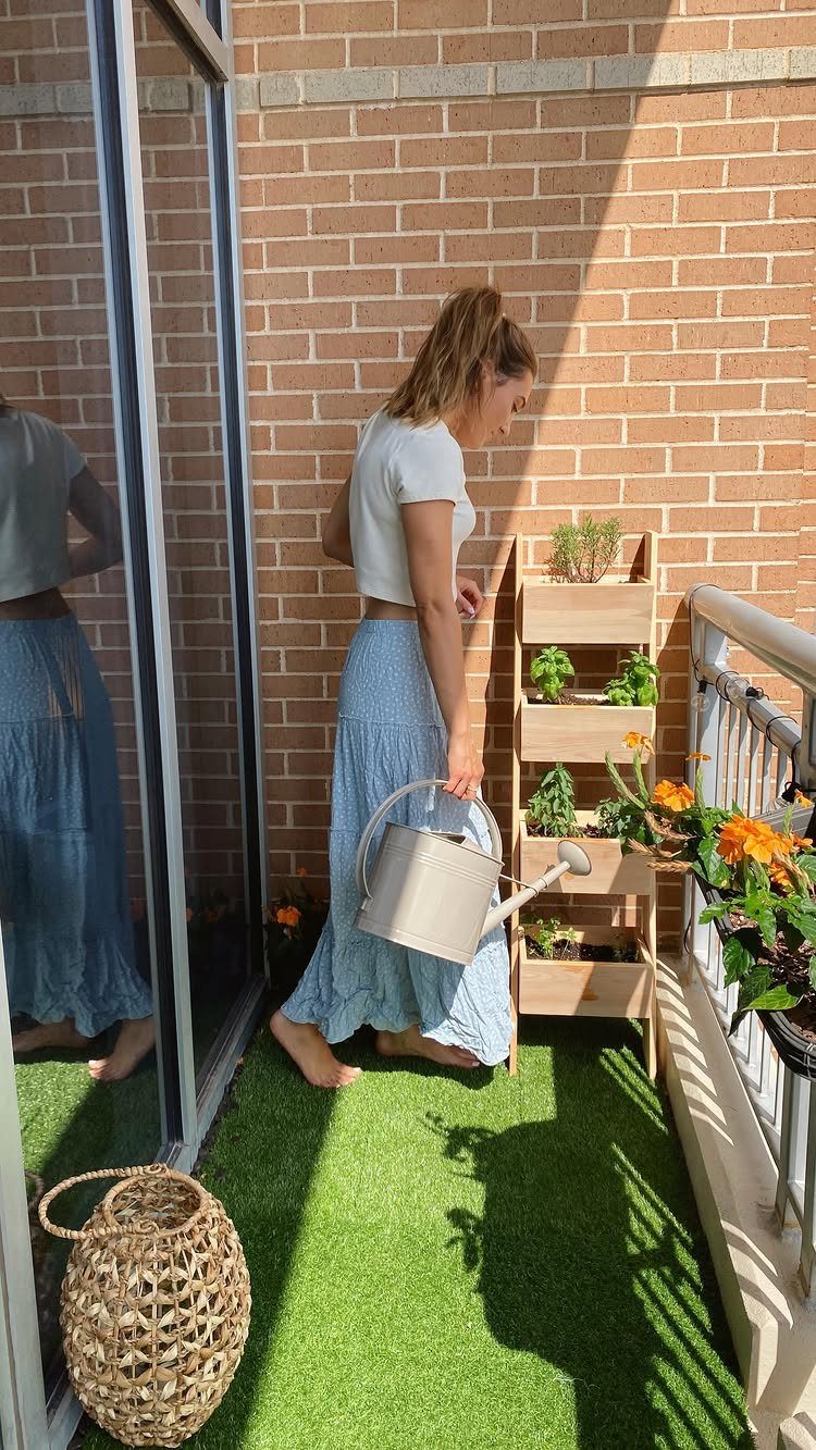 A woman watering small green herbs growing in a wooden tiered plant stand on a balcony.