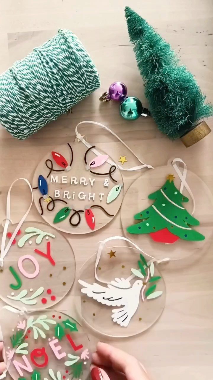 Clear acrylic Christmas ornaments with colorful festive designs and text on a light wooden surface.