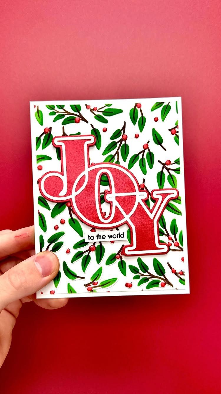 A hand holding a handmade Christmas card with red joy lettering and holly berry patterns.