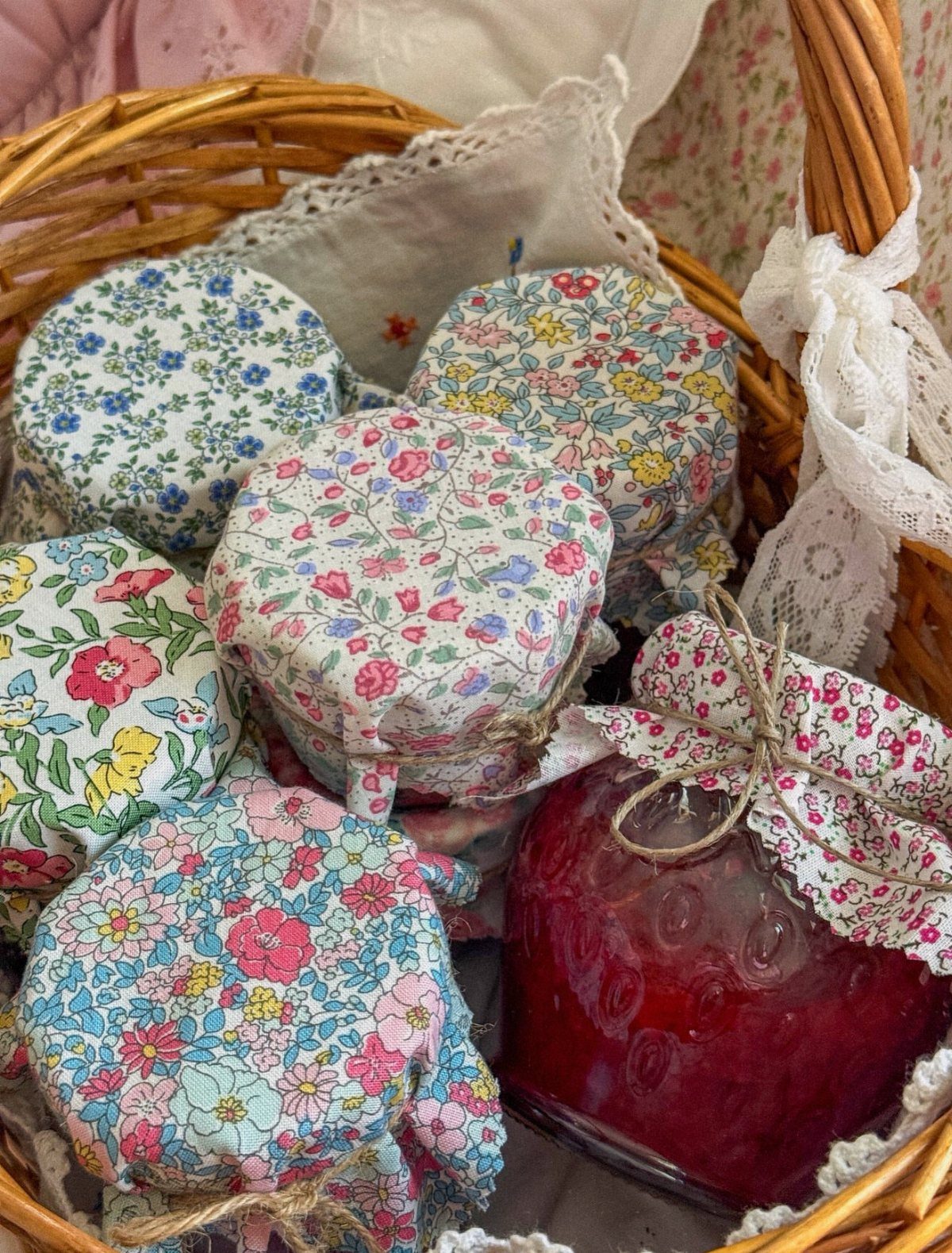 Glass jars of red jam with floral fabric covers inside a wicker basket.