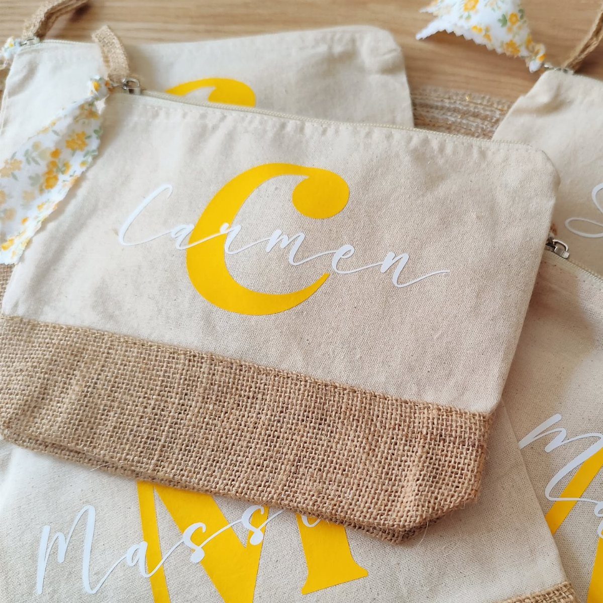 Monogrammed canvas pouches with yellow letters and white cursive names on a wooden surface.