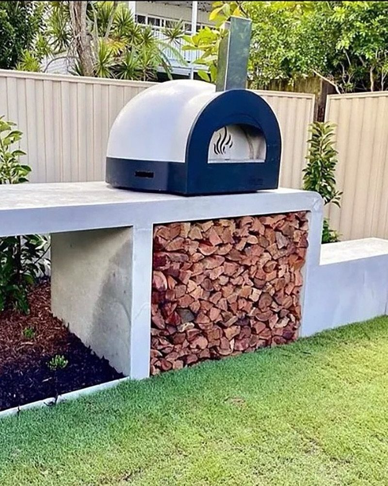A white and black dome pizza oven sitting on a concrete counter with firewood underneath.