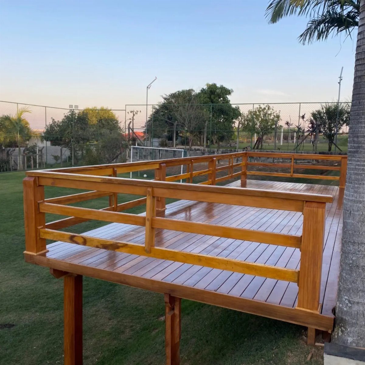 Decks de Madeira: 7 Ideas for Outdoor Living Spaces 7 A polished wooden deck with railings built on a green lawn next to a palm tree.