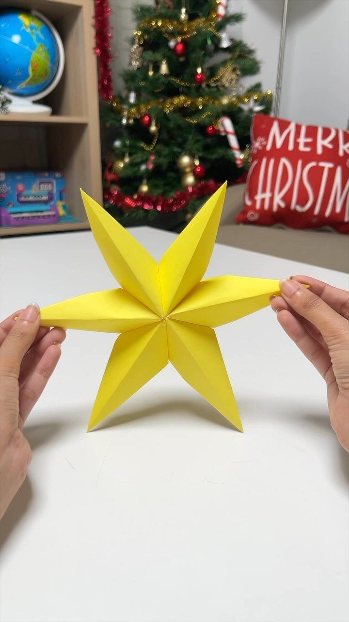 Hands holding a bright yellow 3D origami star on a white table with a Christmas tree background.