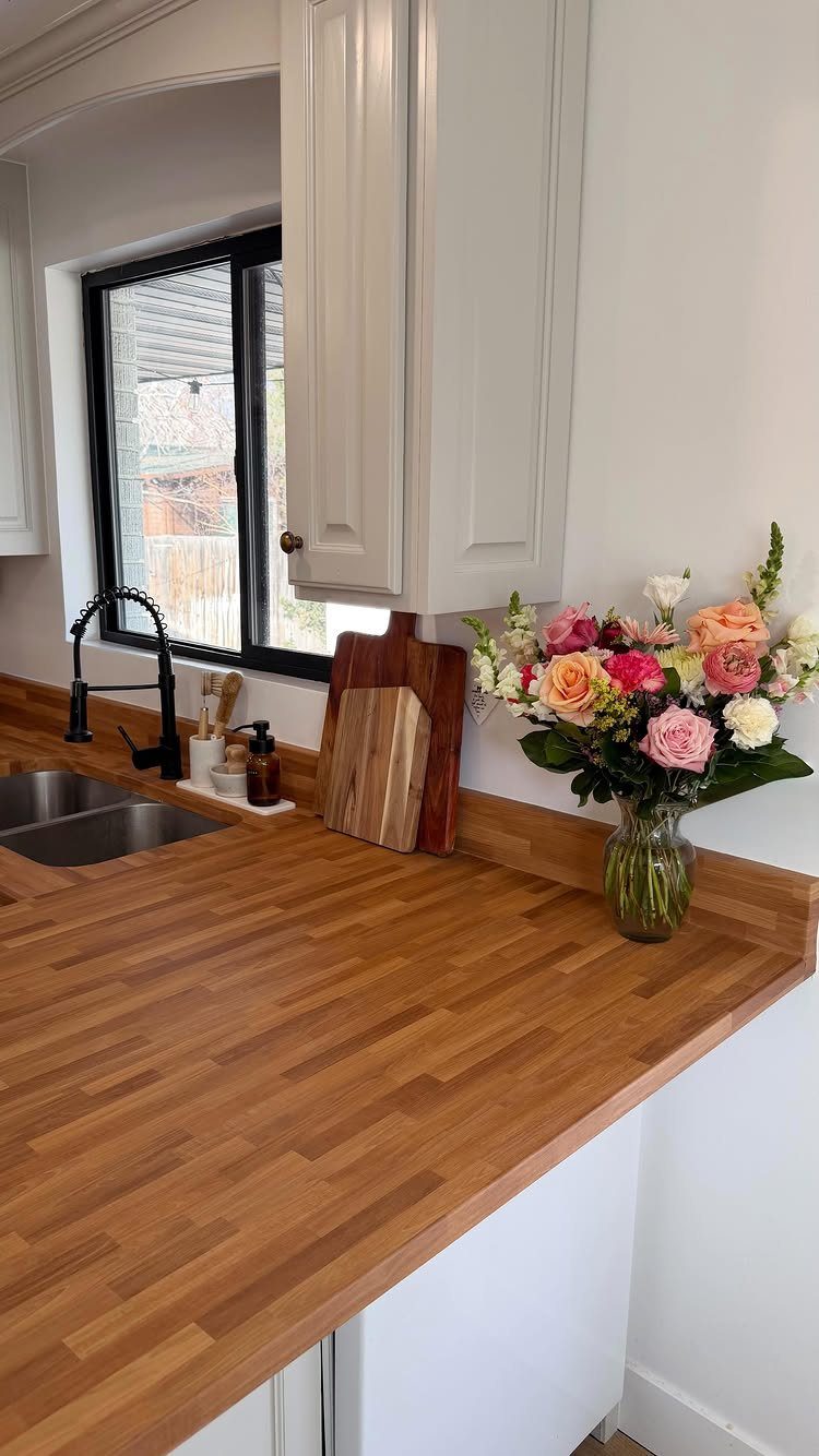 Butcher block wooden countertop with a double sink, black faucet, and a vase of flowers in a kitchen.