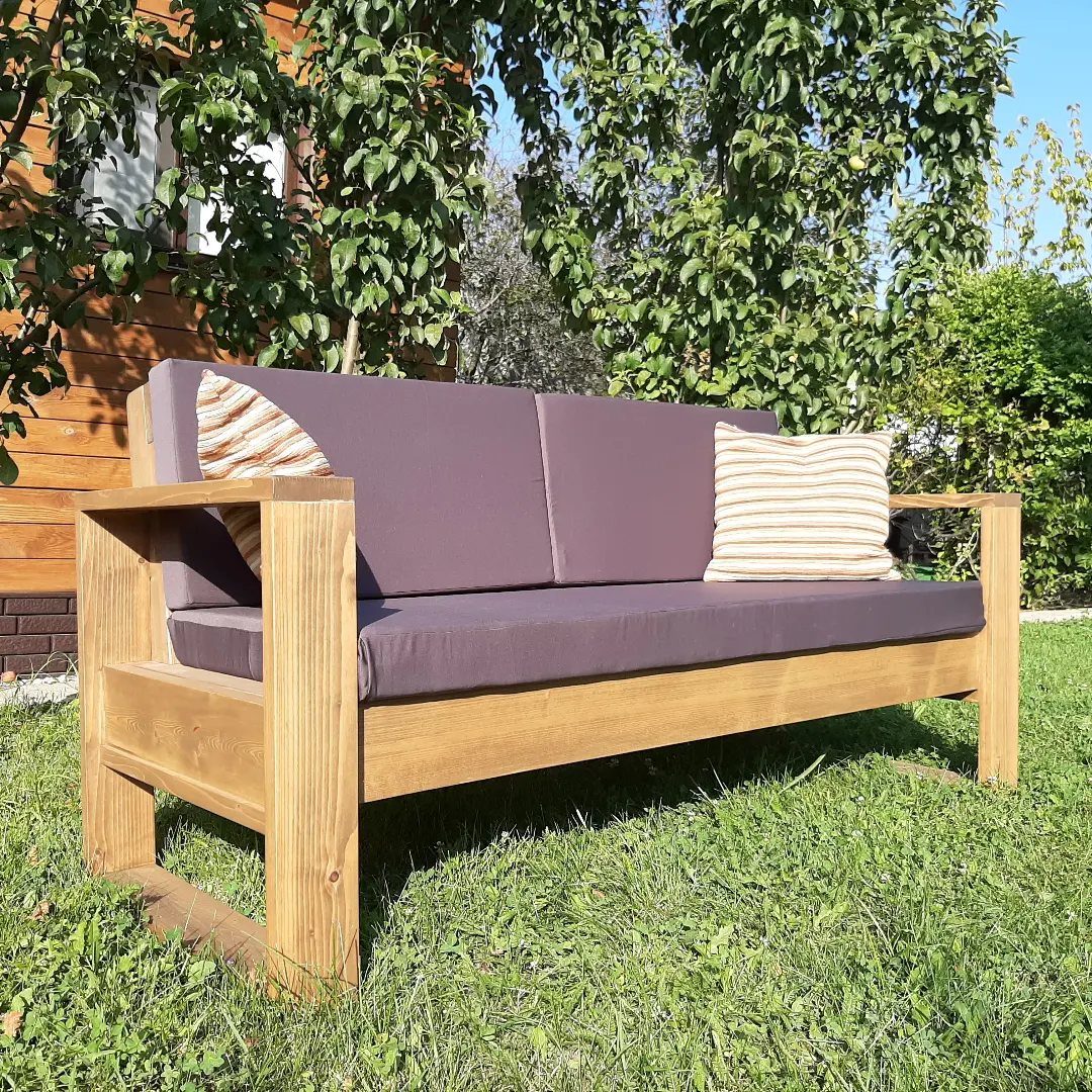 DIY Patio Couch: 8 Projects to Build for Any Budget 2 A modern wooden outdoor couch with purple cushions resting on a green grass lawn.