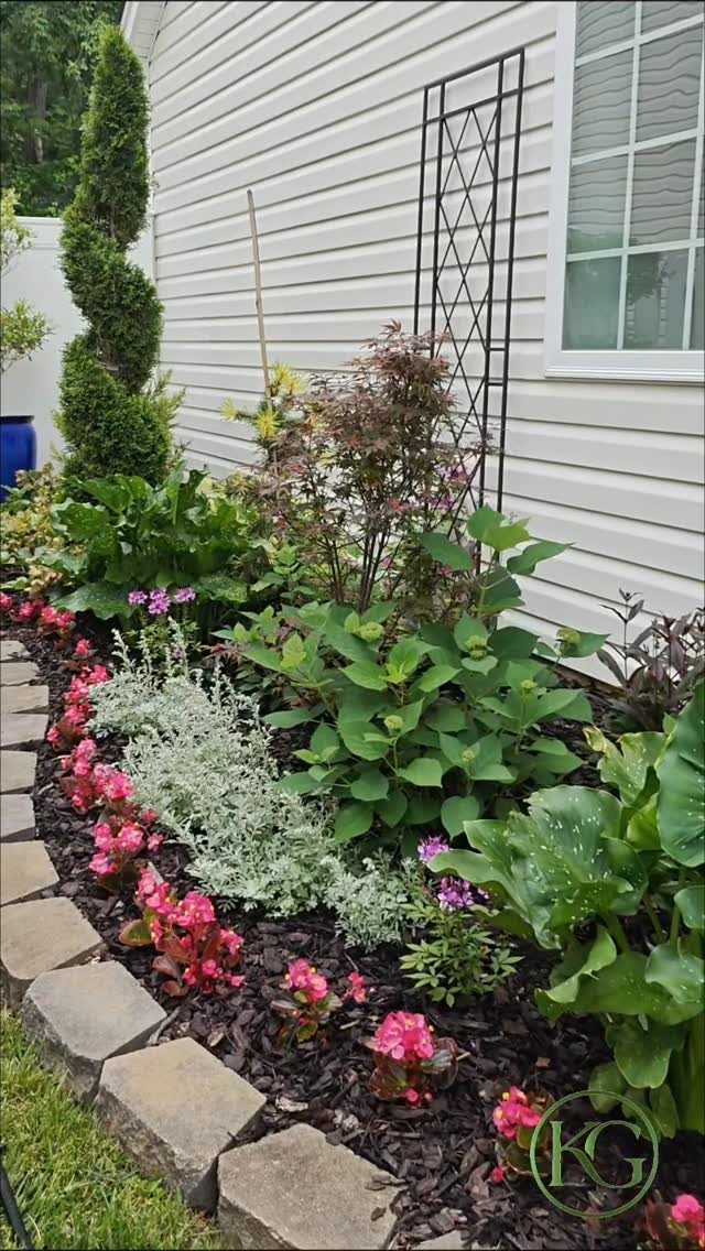 A lush garden bed with pink flowers and green shrubs planted along a white house wall.