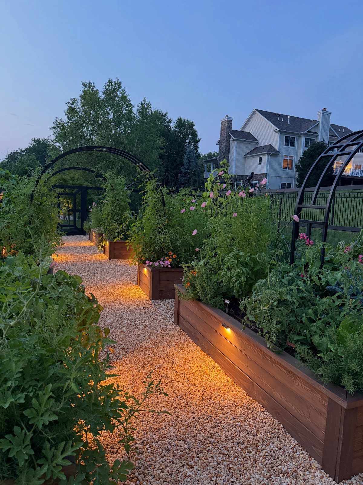 Warm lights glow from wooden raised garden beds lining a gravel path at twilight.