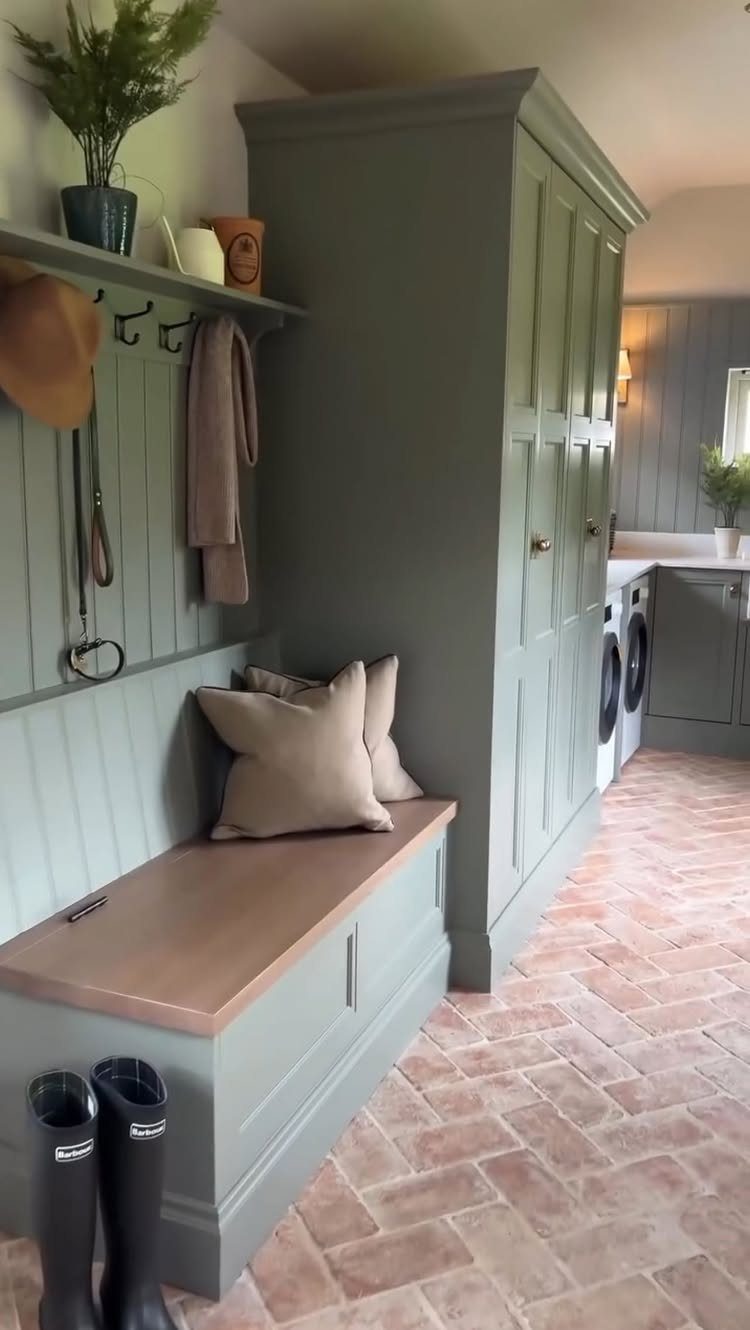 Sage green mudroom bench with wooden seat, pillows, and tall wardrobe on tile flooring.