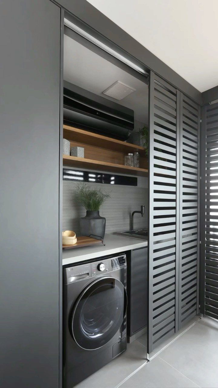 A modern compact utility nook with a washing machine, sink, and wooden shelves behind grey slatted doors.