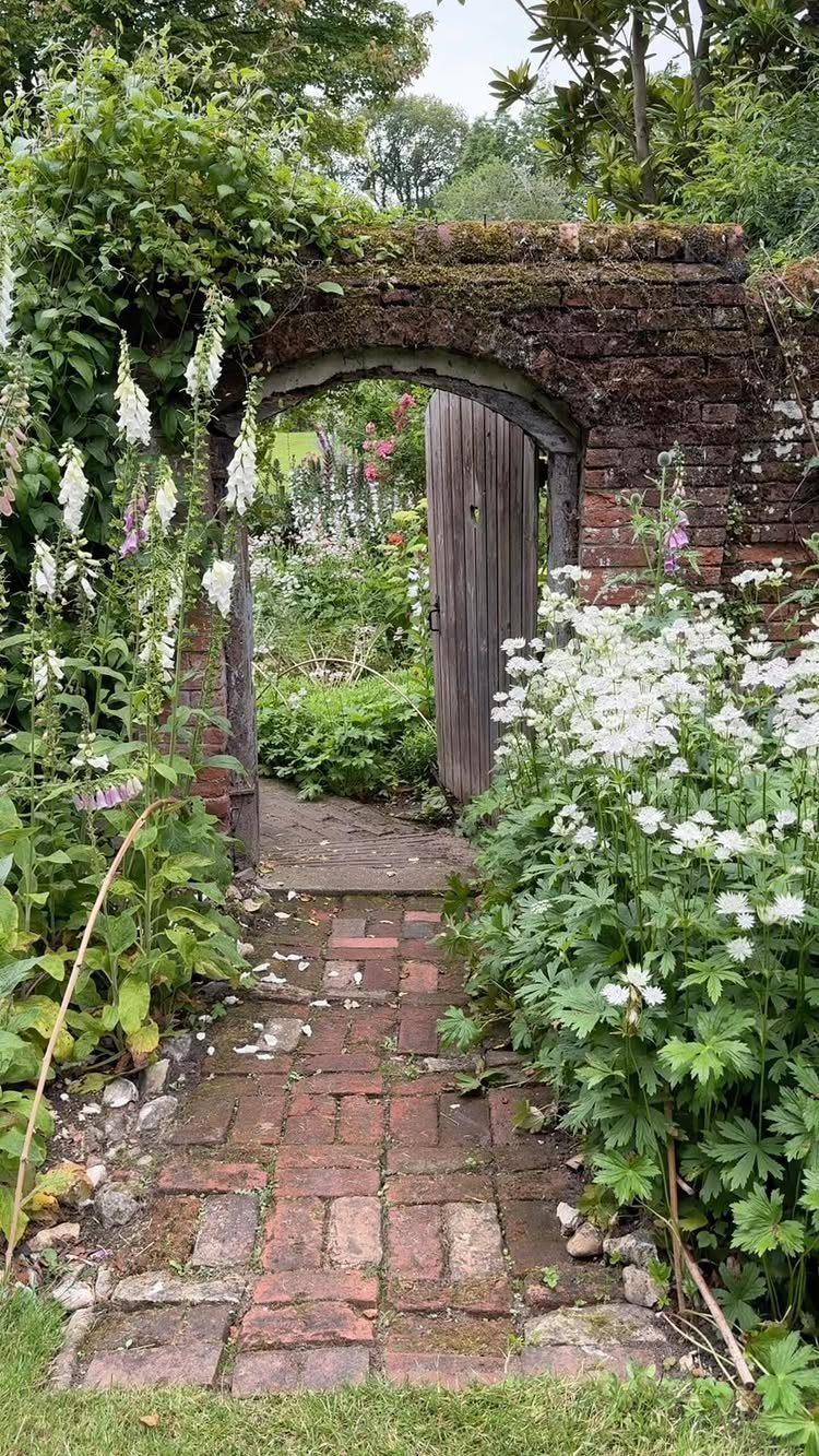 A brick archway with an open wooden gate leading into a lush, flowering garden.