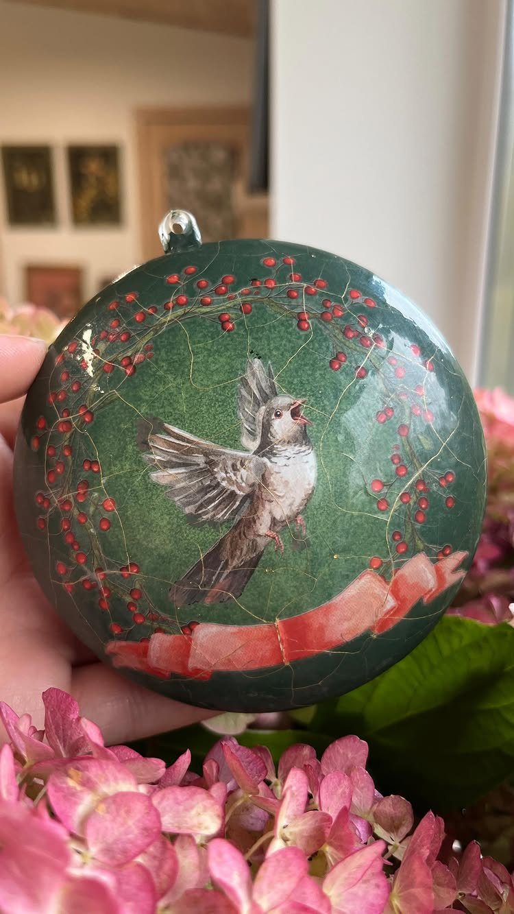 A hand holding a green round ornament with a bird and red berry design.