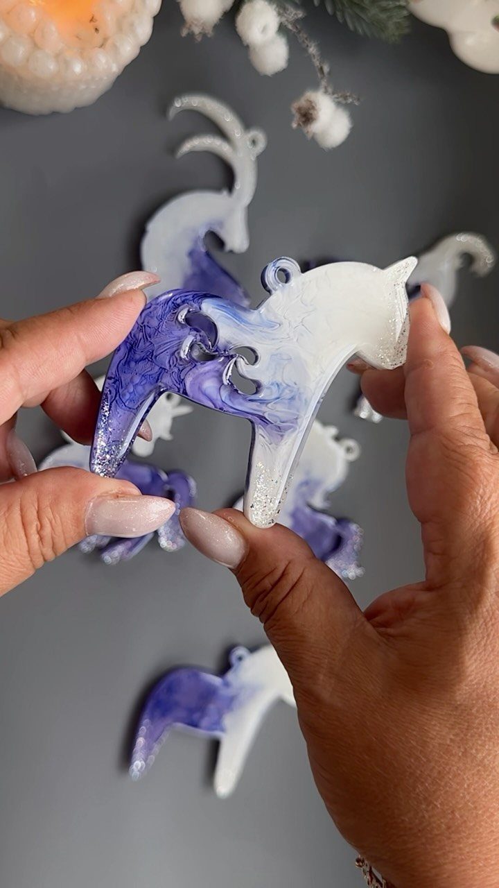 Hands holding a white and indigo marbled reindeer shaped Christmas ornament against a grey background.